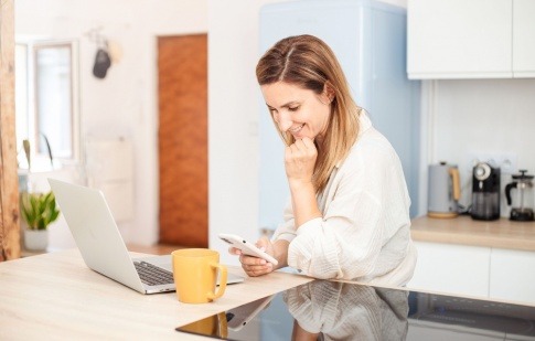 Frau mit Smartphone und Notebook sitzt in Küche mit Elektrogeräten