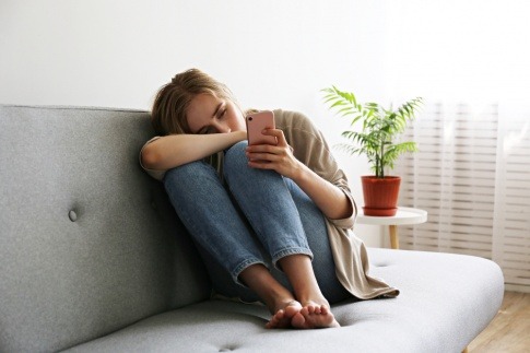 Eine Frau sitzt in zusammengezogener Körperhaltung auf der Couch und schaut traurig in ihr Handy