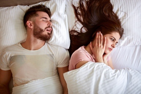 Frau im Bett hält sich die Ohren zu, weil der Partner schnarcht