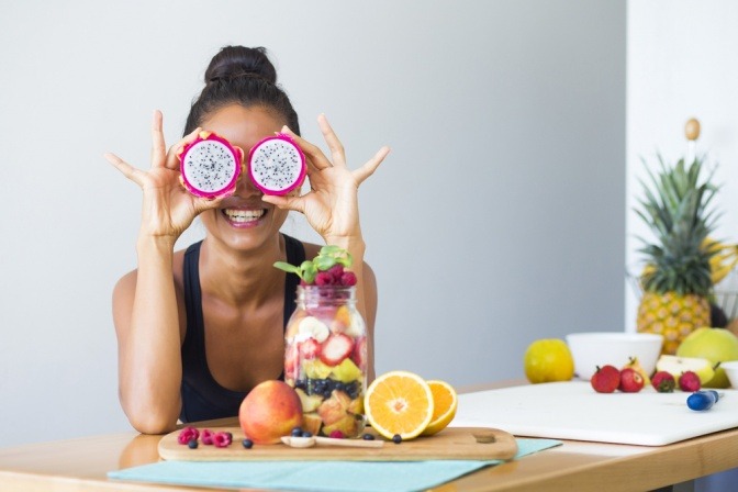 Eine Frau ist mit einem tropischen Fruchtsalat abgebildet und sie verdeckt sich die Augen mit einer Dragon Fruit.