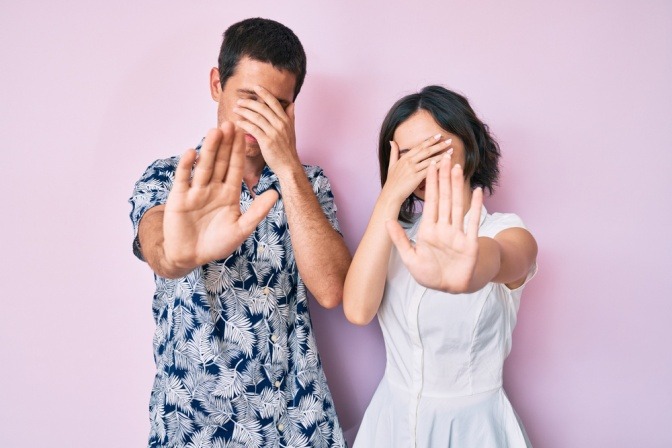 Das Thema Angst vor Beziehung, dargestellt an einem Foto eines Paares, bei dem sich beide Partner eine Hand vors Gesicht halten und mit der anderen Hand die Stopp-Geste machen.  
