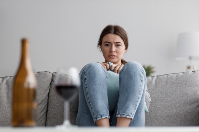Eine traurige junge Frau sitzt allein auf dem Sofa, vor ihr steht eine halbleere Flasche Wein auf dem Tisch.