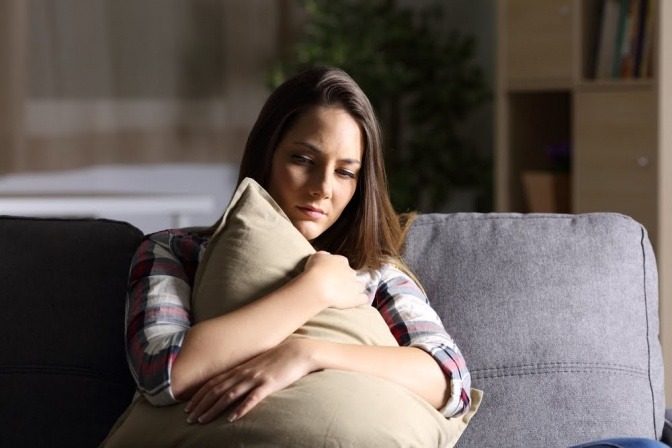 Eine Frau sitzt in einem dunklen Zimmer auf einem Sofa, umarmt ein Kissen und wirkt betrübt, nachdenklich und unsicher.