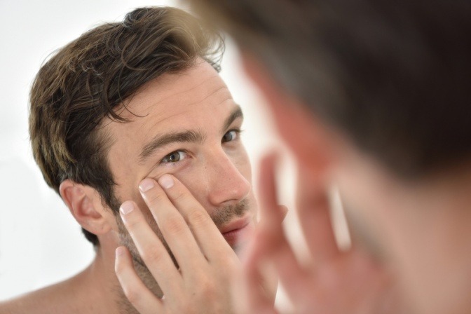 Ein Mann betrachtet im Spiegel seine Haut um die Augen