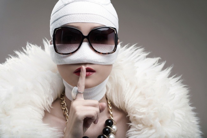 Eine im Gesicht bandagierte Frau zeigt die bekannte "Ruhig jetzt"-Geste mit einem durchgestreckten Indexfinger vor ihrem Mund.