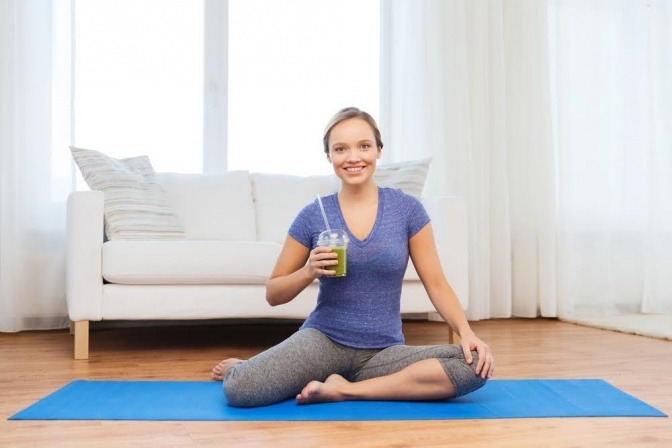 Eine junge Frau sitzt in ihrem Wohnzimmer im Sport-Outfit auf einer Iso-Matte und blickt lächelnd in die Kamera. Neben ihr steht ein Becher mit einem grünen Smoothie.