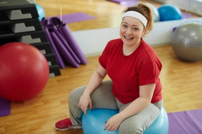 Eine Frau sitzt in einem Sportraum auf einem Gymnastikball und blickt zufrieden in die Kamera.