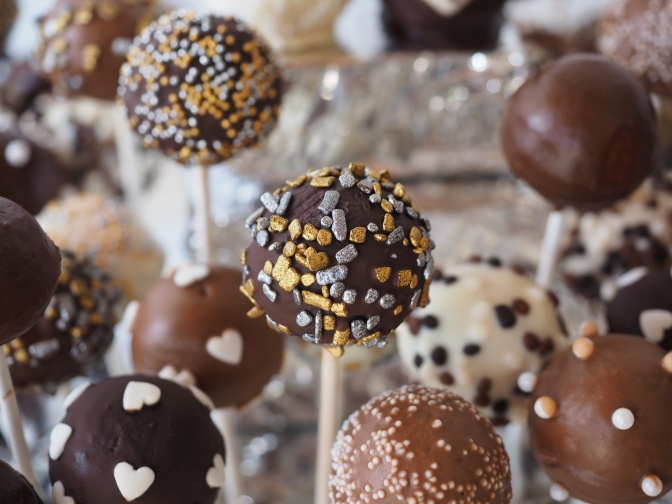 Cake Pops sind in einer Großaufnahme abgebildet. Die meisten sind mit dunkler Schokolade überzogen, im Hintergrund sieht man aber auch weiße.