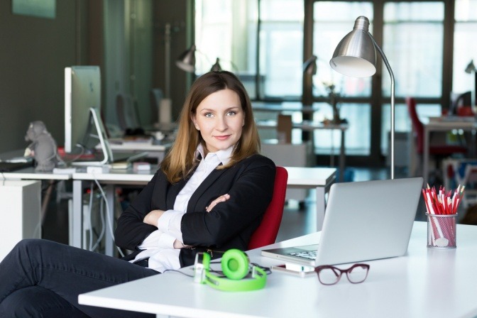 Frau in Businessoutfit sitzt im Cafe und blickt fordernd und selbstbewusst in die Kamera.