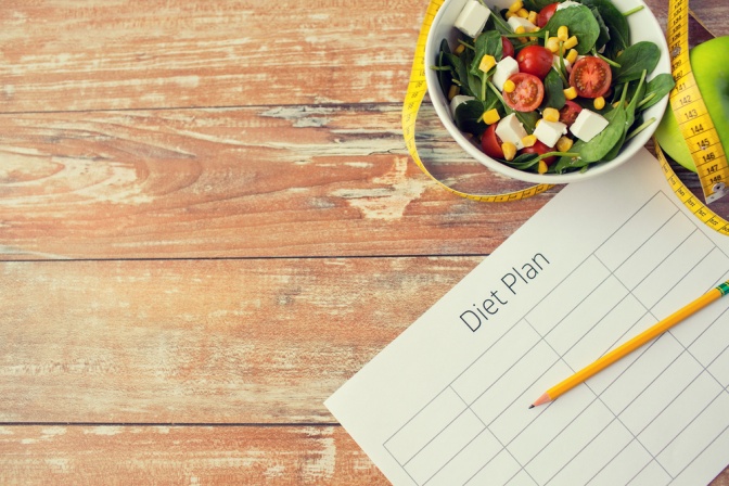 Auf einem hölzernen Untergrund liegen ein Zettel mit Stift und eine kleine Schüssel Salat. Auf dem Zettel steht geschrieben: "Diätplan".
