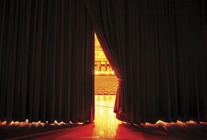 Ein Theatervorhang, der leicht geöffnet ist und einen Blick in den Zuschauerraum freigibt. 