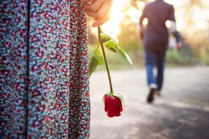 Eine Frau hält eine Rose in einem am Körper anliegenden Arm, während Ihr vermeintlicher Partner gerade von ihr weg geht.