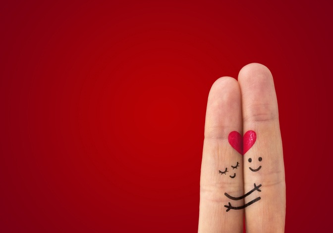 Wir sehen zwei Finger einer Hand, Zeige- und Mittelfinger, die ganz eng aneinander liegen. Ein romantisches Smiley ist mit roter Farbe auf die Fingerkuppen gemalt.  