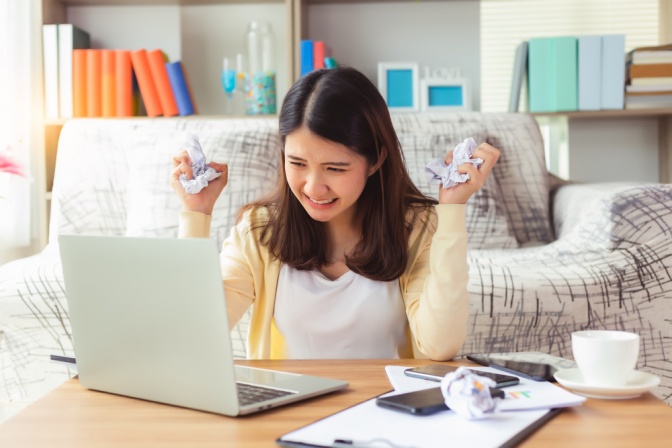 Junge Frau zerknüllt Papier und wirkt frustriert, während sie vor dem Computer sitzt. 