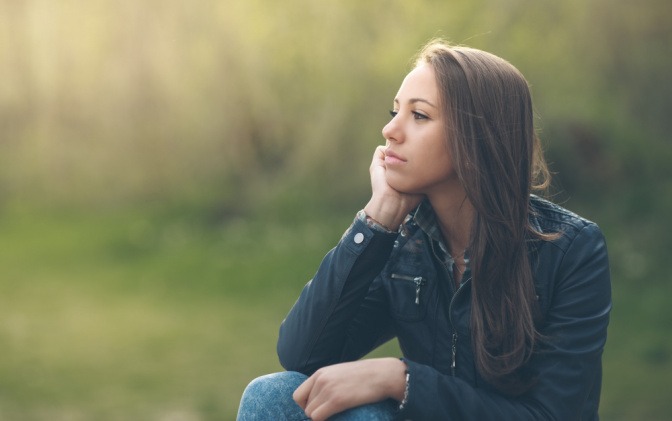 Eine junge Frau sitzt in einem Park und blickt nachdenklich in die Ferne.