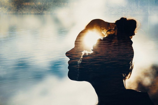 Die seitliche Schattensilhoeutte einer Frau, die an einem See sitzt. Ihr Körper ist transparent. Es wirkt, als sei die Energie ihres Körpers mit der spirituellen Energie der Umgebung verknüpft. 