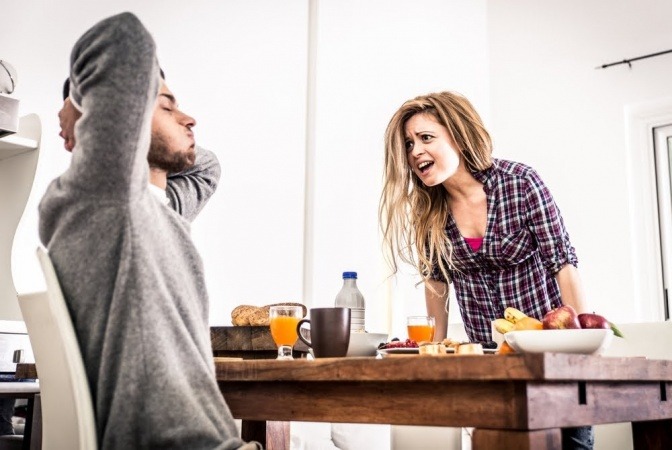 Ein Paar streitet sich, der Mann sitzt am Tisch und macht einen resignierten Eindruck, die Frau steht gegenüber und wirkt aufgeregt und verärgert.