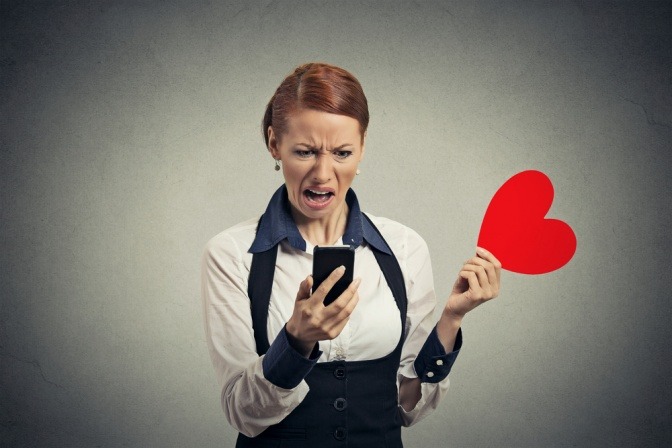 Eine unzufrieden und schockiert wirkende Frau liest Nachrichten auf ihrem Smartphone und wirft ein rotes Plüschherz weg. 