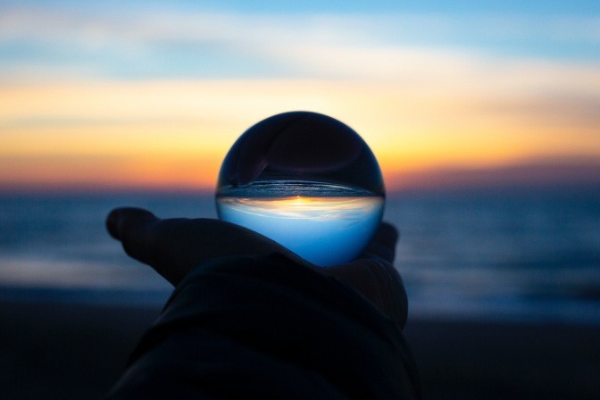 Jemand hält eine große transparente Glaskugel vor sich in den Händen, in der sich das Bild einer untergehenden Sonne über dem Meer spiegelt.