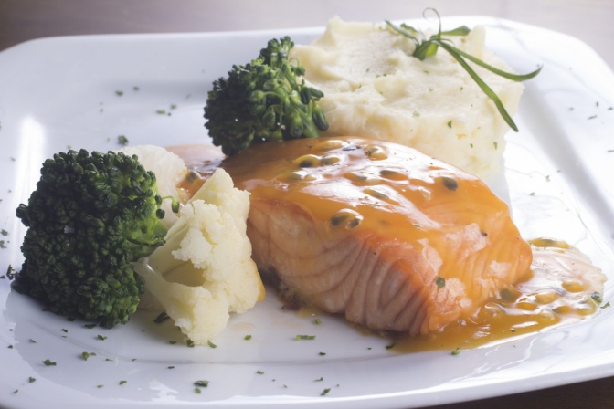 Ein weißes Teller, auf dem Lachs und Gemüse sind, ist abgebildet. Der Lachs ist mit einer Sauce übergossen.