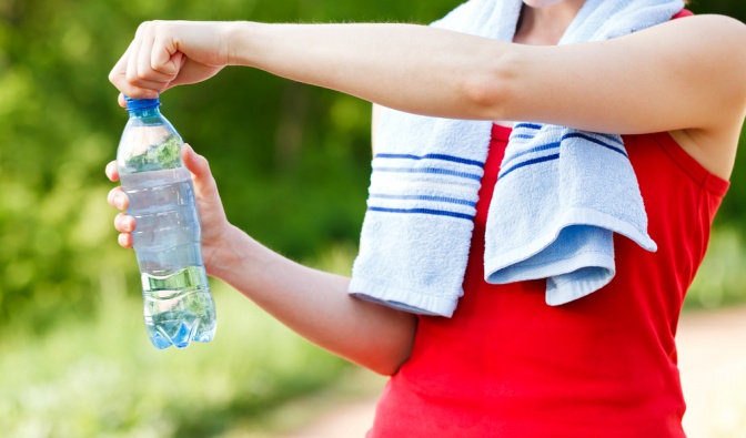 Eine Frau in sportlicher Bekleidung öffnet eine Wasserflasche. Um ihre Schultern hat sie ein Handtuch gelegt. Ihr Top ist rot. Auf dem Foto ist nur ihr Brust- und Bauchbereich abgebildet, das Gesicht sieht man nicht.