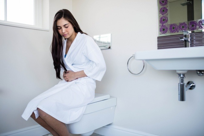 Eine Frau sitzt im Bademantel auf der Toilette und hält den Bauch