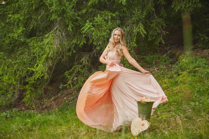 Eine Frau mit rosa Kleid tanzt neben einem Herz aus Zirbenholz im Wald