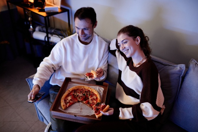 Freundschaft nach Beziehung - Mann und Frau sitzen auf Sofa, teilen sich eine Pizza und schauen TV.