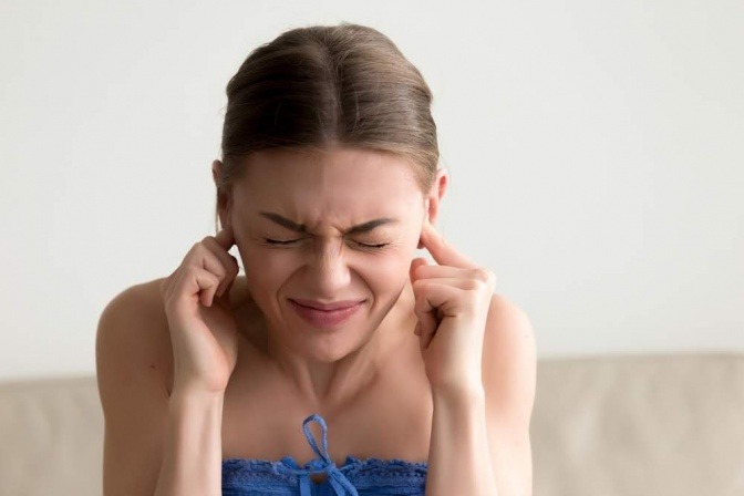 Eine Frau hält sich die Ohren zu und macht ein schmerzverzerrtes Gesicht.