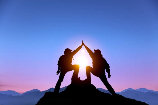 Ein Paar hat gemeinsam den Gipfel eines Bergs erklommen und gibt sich nun an der Spitze ein High Five, während die Sonne im Hintergrund untergeht.