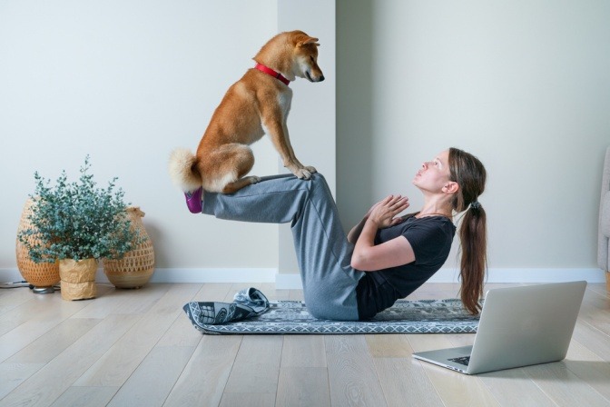 Frau macht Yoga und balanciert dabei einen Hund auf ihren Beinen.