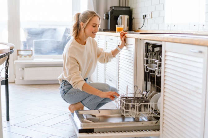 Lächelnde junge Frau beim Einräumen der Geschirrspülmaschine.