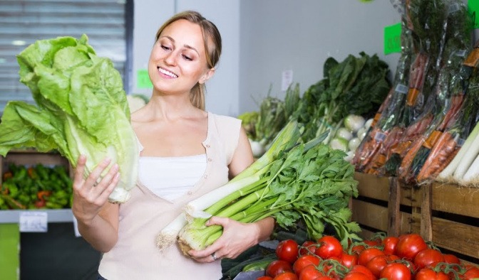 Eine Frau steht in ihrer Küche und hält diverse gesunde Lebensmittel wie Salate und anderes Gemüse in den Händen, mit denen sie gleich gesund kochen wird.