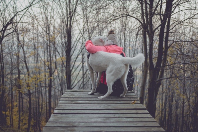 Eine Frau sitzt im Winter an einem Steg und umarmt ihren Hund, der neben ihr sitzt.