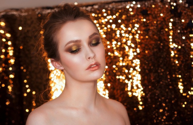 Eine Frau steht vor einem goldenen Hintergrund und trägt goldenes Glitzer-Makeup
