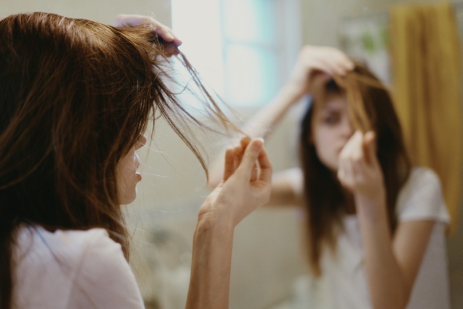 Frustrierte junge Frau inspiziert ihre spröden Haare vor dem Badezimmerspiegel.