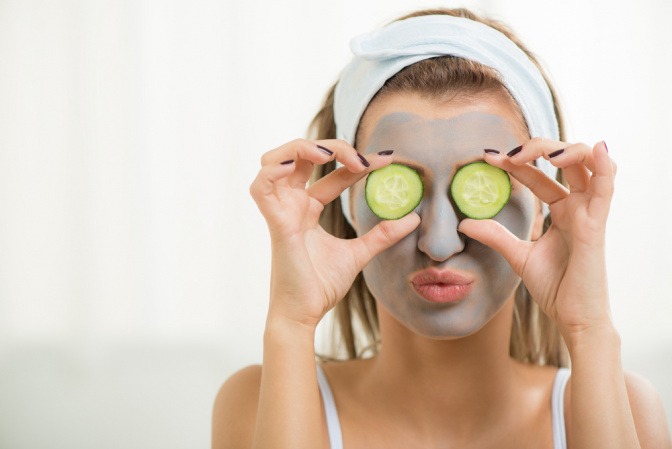 Eine Frau hält Gurkenscheiben vor ihre Augen und trägt eine Gesichtsmaske