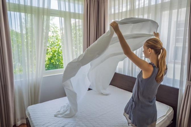 Frau zieht Bettwäsche ab und lüftet Schlafzimmer gegen Hausstaubmilben