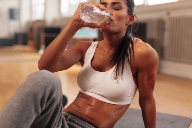 Eine stark verschwitzte Frau in einem Trainingsraum macht zwischen HIIT Übungen eine Pause und trinkt Wasser aus einer Flasche.