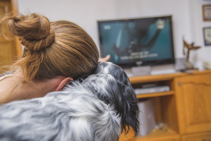 Frau sitzt mit ihrem Hund auf dem Sofa und schaut etwas im TV.