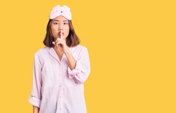 Eine Frau im Pyjama hält sich den Finger vor den Mund und macht die typische "Psst"-Geste.