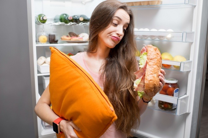 Frau steht abends vor dem geöffneten Kühlschrank. In der einen Hand hat sie ein Kissen, in der anderen ein riesiges belegtes Brot.