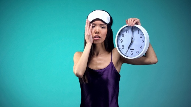 Innere Uhr umstellen - müde wirkende Frau im Nachthemd hält eine Wanduhr hoch.