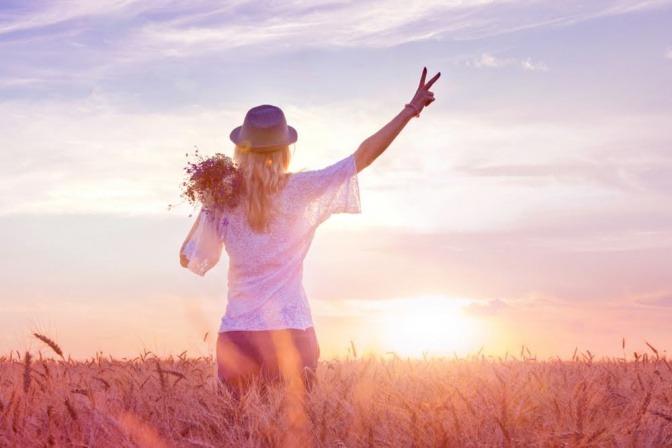 Eine Frau steht bei Sonnenuntergang in einem Blumenfeld und macht mit der rechten erhobenen Hand das Victory-Zeichen.