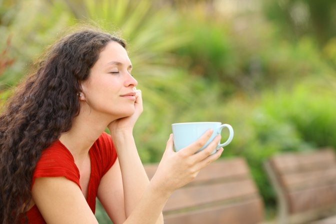 Entspannte Frau sitzt in einem Feld mit hohem Gras und trinkt eine Tasse Tee.