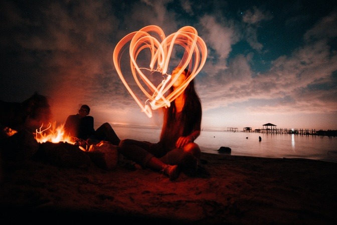 Eine Frau sitzt am Strand und malt mit einem Laserpointer Herzsymbole in die Luft.