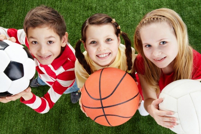 Drei kleine Kinder mit unterschiedlichen Sportbällen im Arm blicken in freudiger Erwartung einer anstehenden Spielstunde hoch zu ihren Eltern.