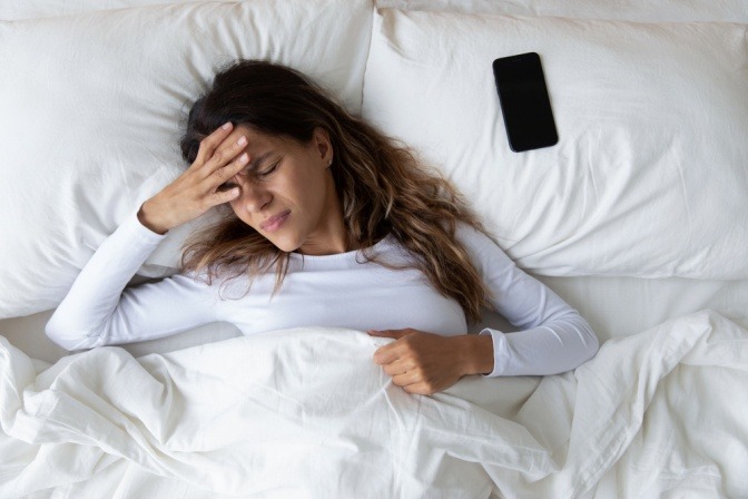 Frau liegt mit Kopfschmerzen im Bett, neben ihr liegt ein Smartphone auf einem Kissen.
