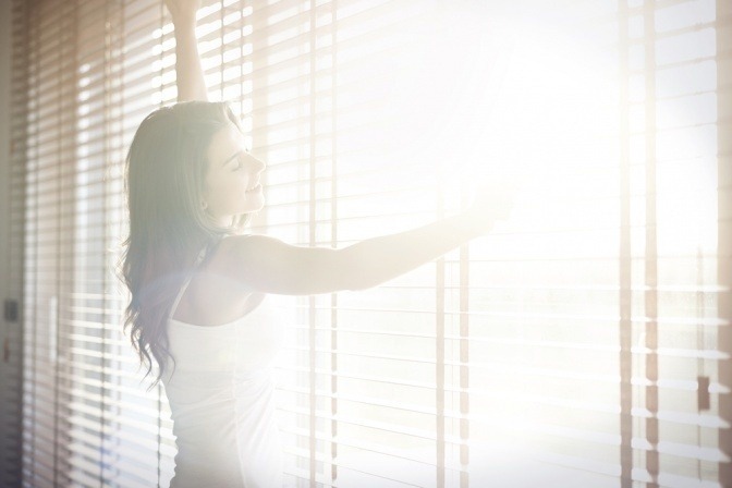 Eine Frau steht vor einem Fenster mit Jalousien, bei dem Licht reinscheint