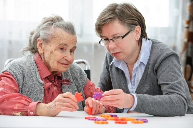Zwei Frauen sitzen gemeinsam am Tisch und lösen ein Puzzle, eine der Frauen ist deutlich älter als die andere. Die Szene könnte in einem Altenheim spielen.